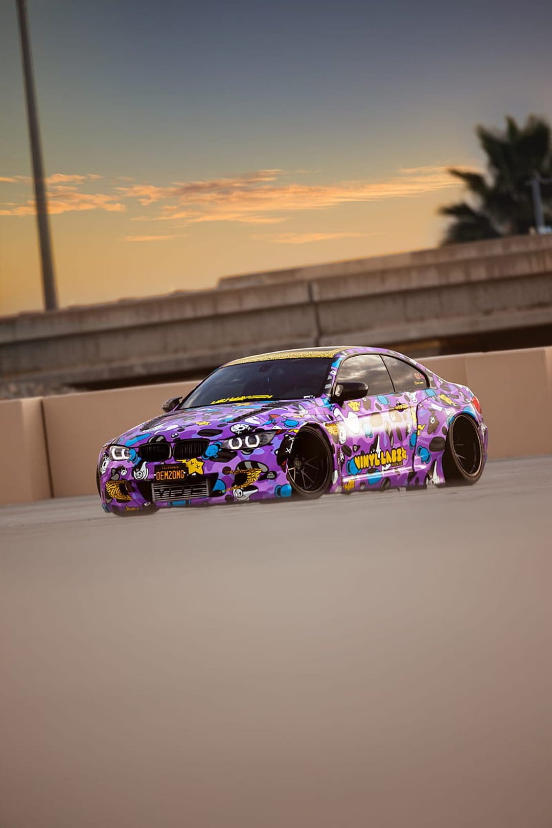 Purple camouflage BMW M3 sports car with black rims on a highway overpass under a warm orange sunset sky.