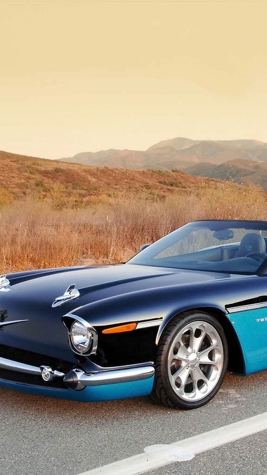 Blue vintage convertible car with chrome accents parked on a mountain road against a backdrop of golden hills.