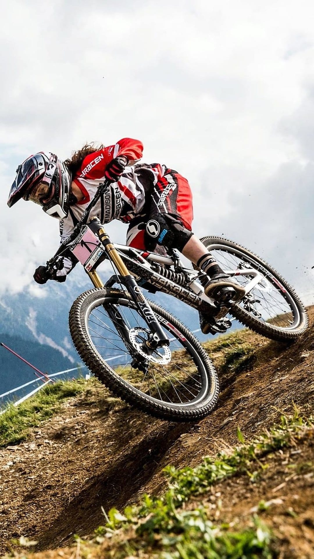 Downhill cyclist in a full-face helmet leans into a sharp dirt turn against a blurred mountain range backdrop.