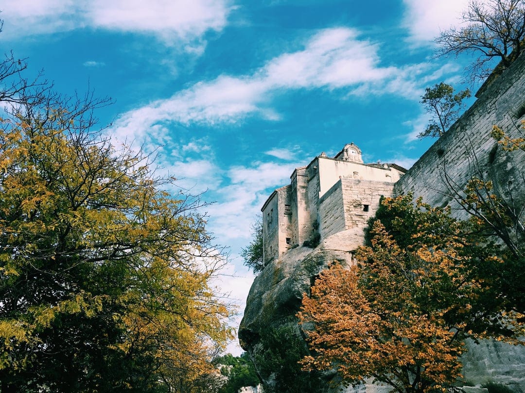 Stone castle atop a rugged cliff face with vibrant orange trees and a cloud-streaked blue sky in the background.