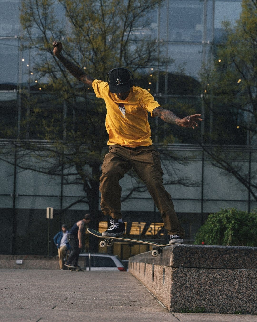 Skateboarder in a yellow shirt grinds a concrete ledge in front of a glass building and green trees in daylight.