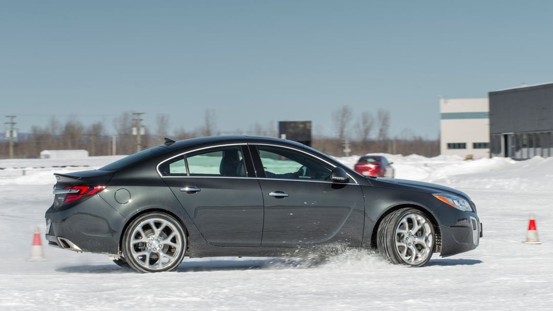 Dark grey sedan drifting on a snowy track with splashing snow and silver wheels under a clear winter sky.