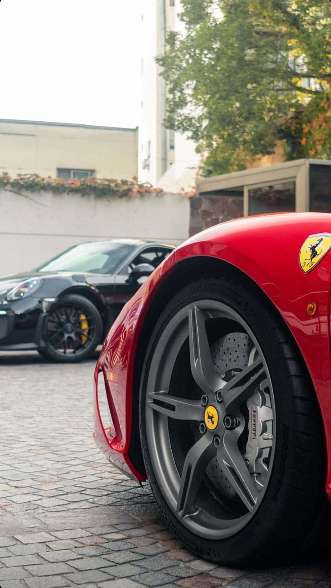 Red Ferrari wheel and fender in focus with a black Porsche parked on cobblestones under bright sunlight.