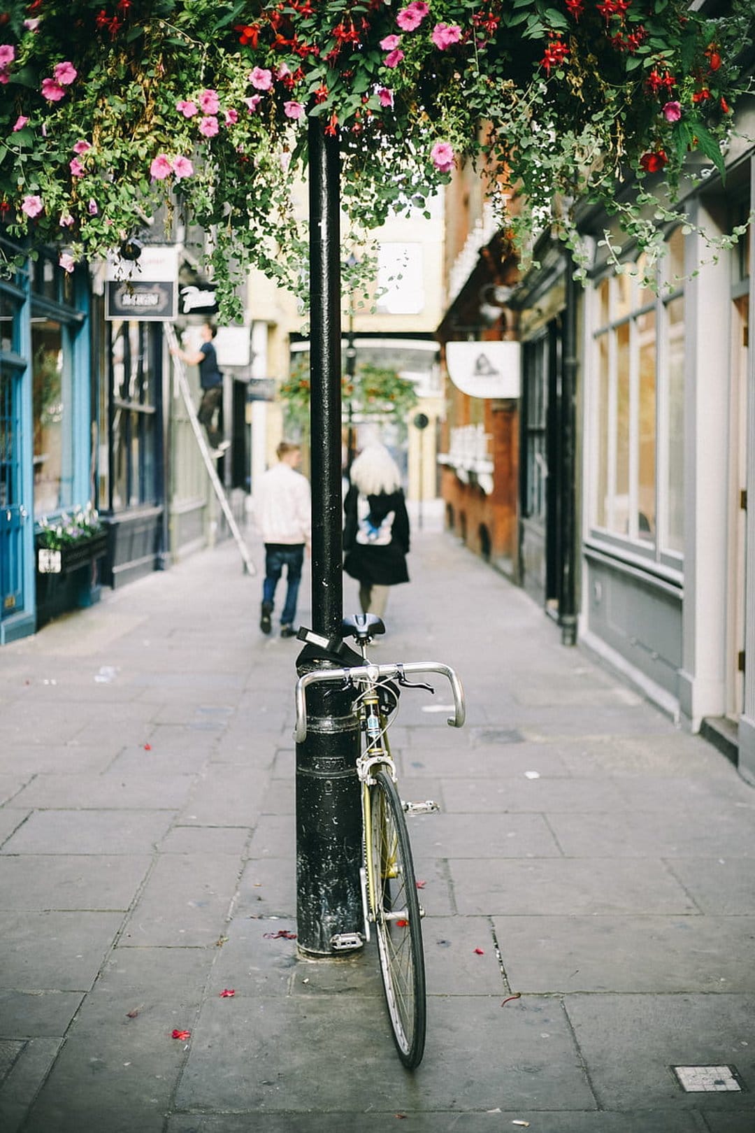 Vintage bicycle by a lamppost covered in pink flowers on a cobblestone street with soft sunlight filtering in.
