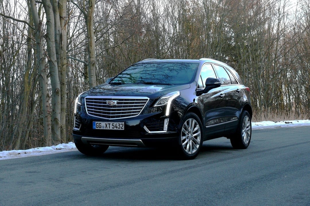Black Cadillac XT5 SUV parked on a paved road through a forest with bare trees and patches of roadside snow.