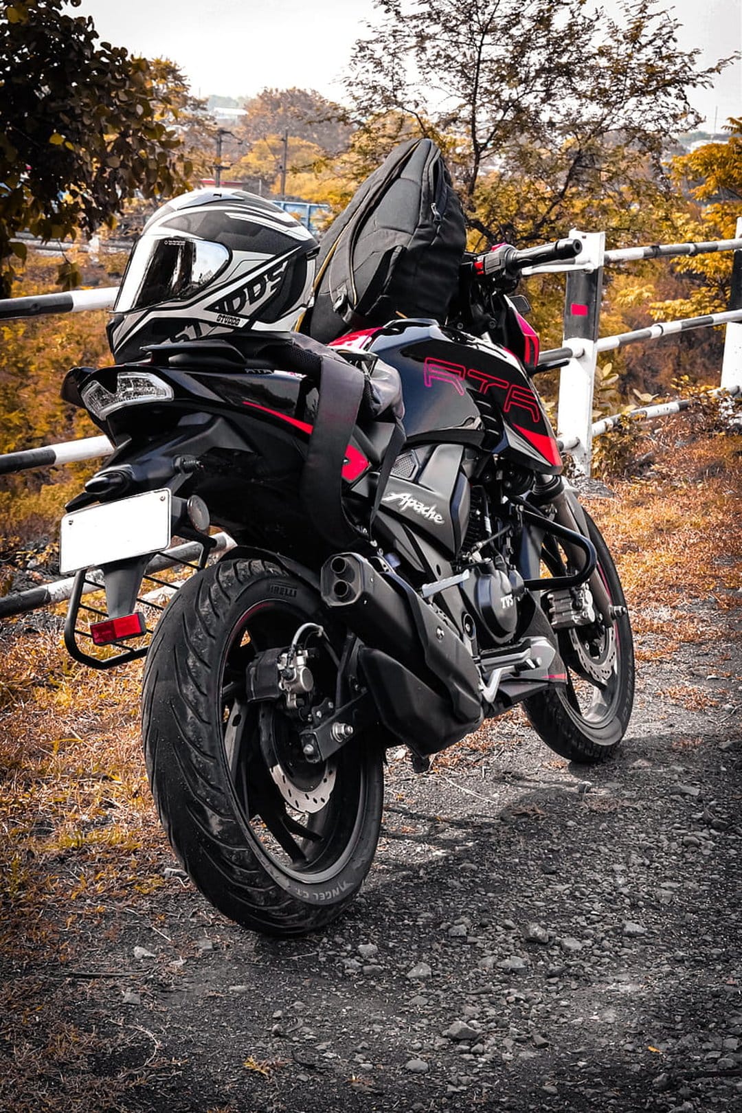 Black TVS Apache motorcycle with a white helmet on the seat and a saddlebag, parked on gravel amidst fall foliage.