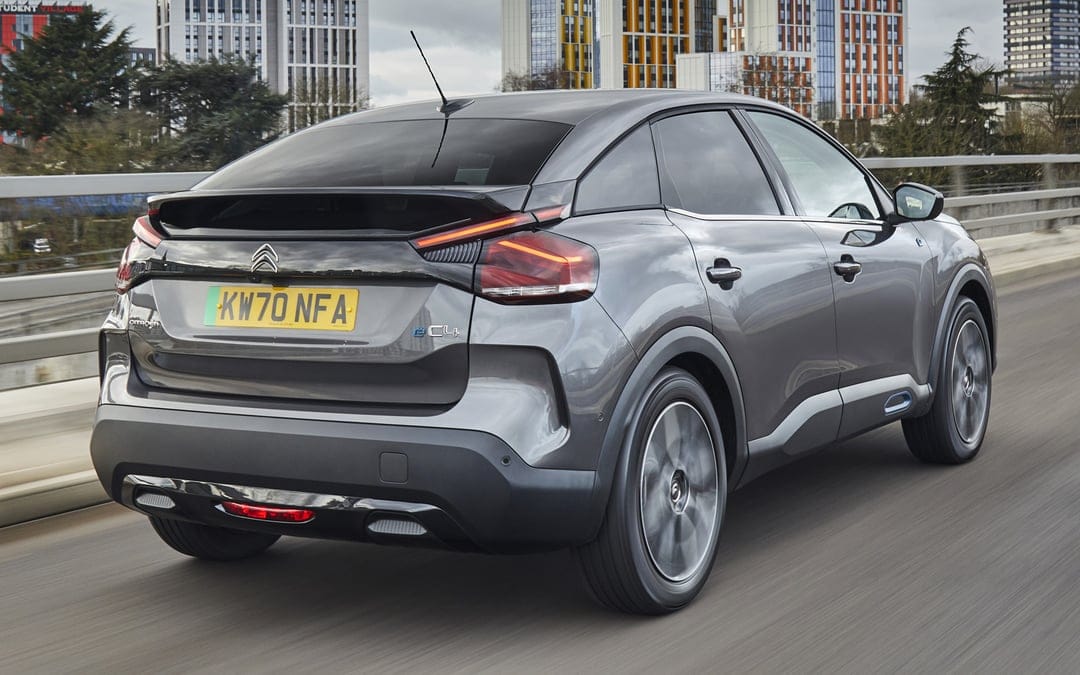 Dark gray Citroen e-C4 electric hatchback driving on a city bridge with tall modern buildings in the background.