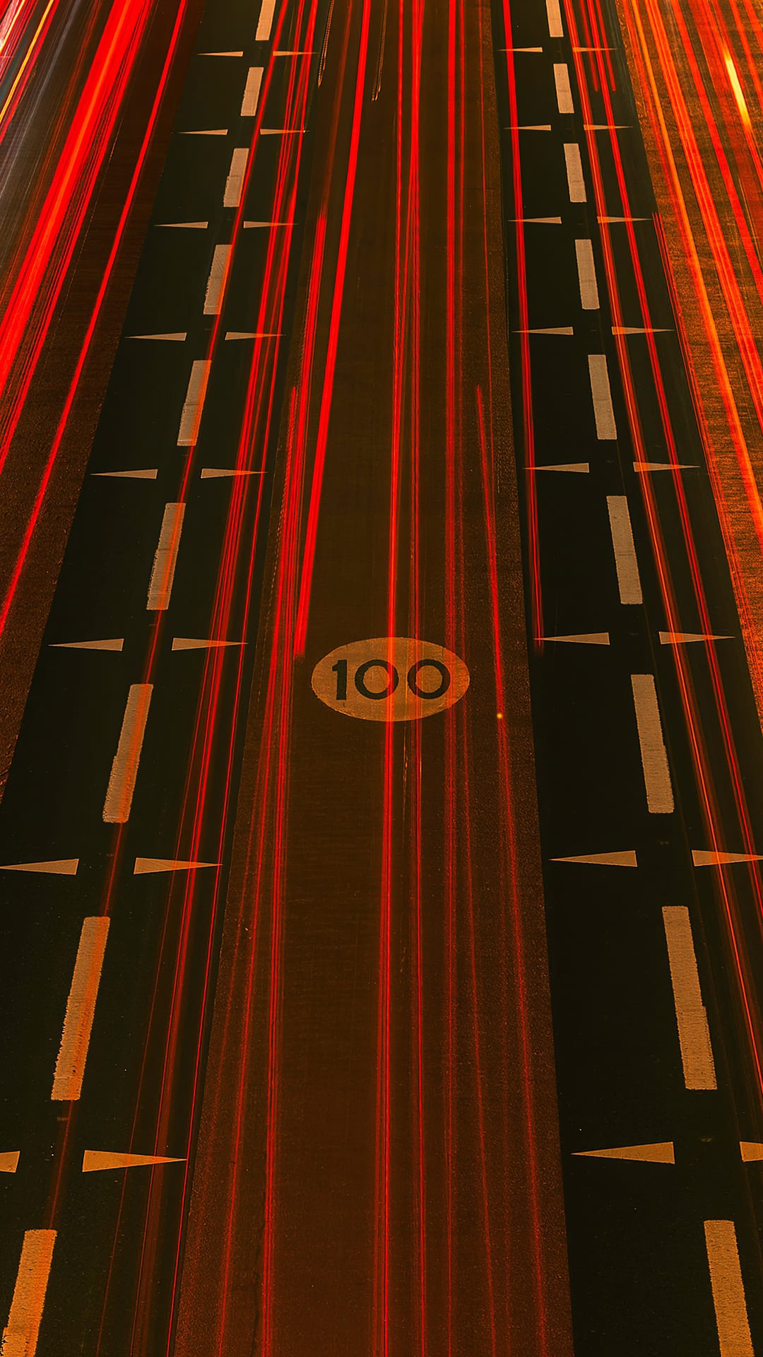 Red light streaks from car taillights flow past a 100 mph speed limit painted on dark highway asphalt at night.