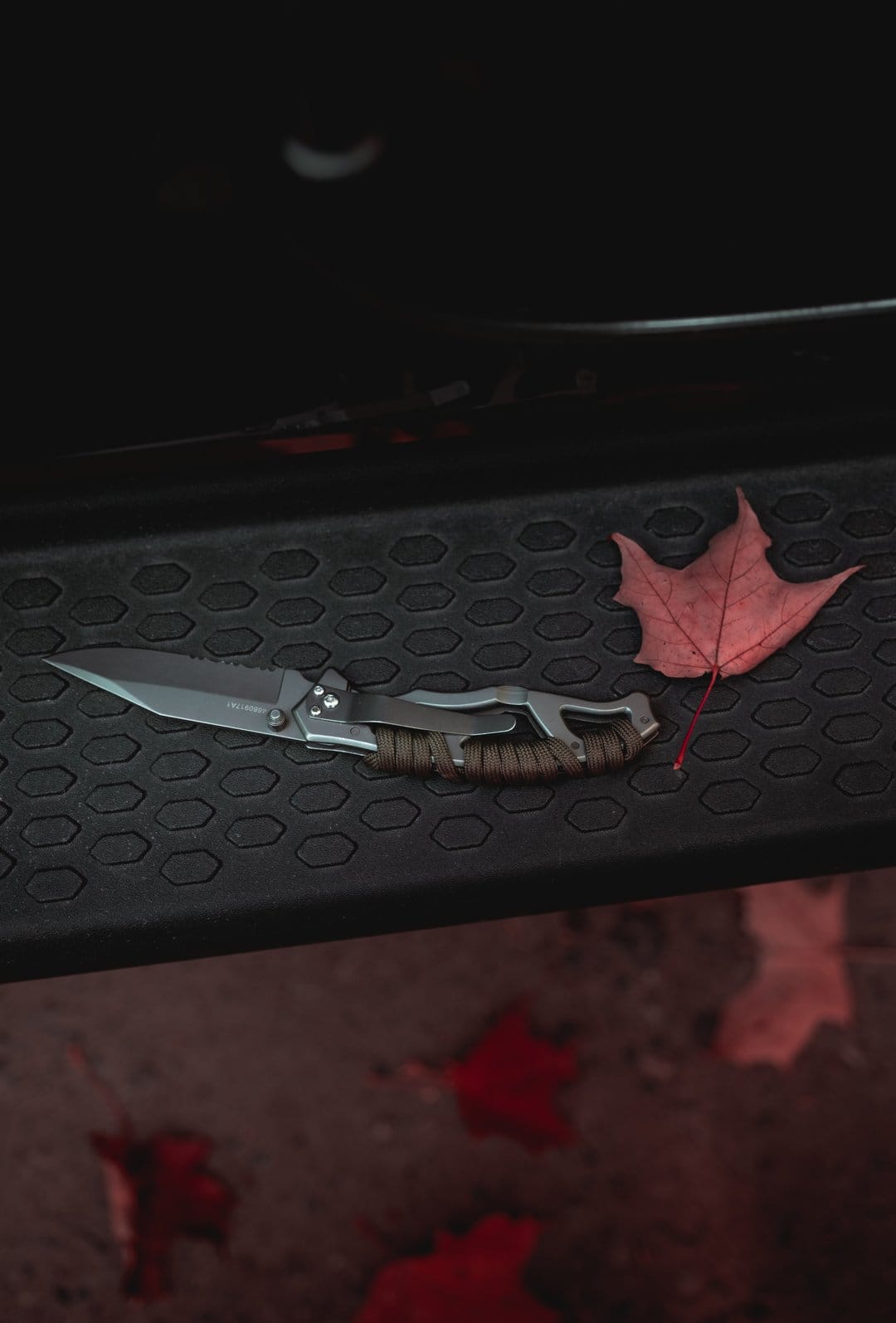 Tactical knife with paracord handle and serrated blade lies next to a red maple leaf on a dark textured background.