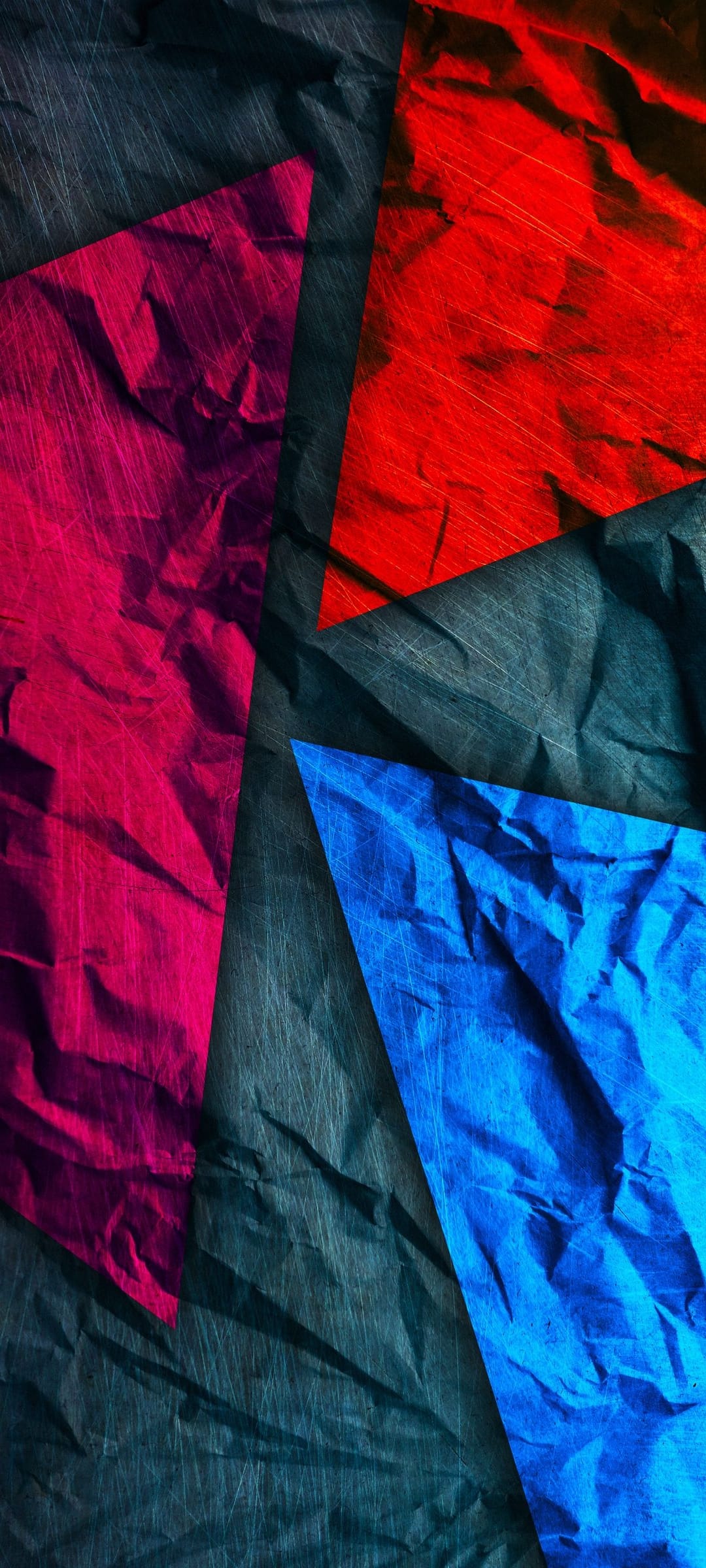 Crushed paper triangles in red, magenta, and blue rest on a dark textured surface with fine scratch marks.