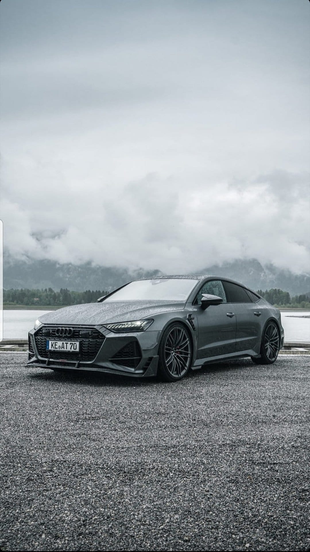 Modified Audi RS7 parked on gravel by a mountain lake under cloudy skies, shown in a vertical mobile orientation.