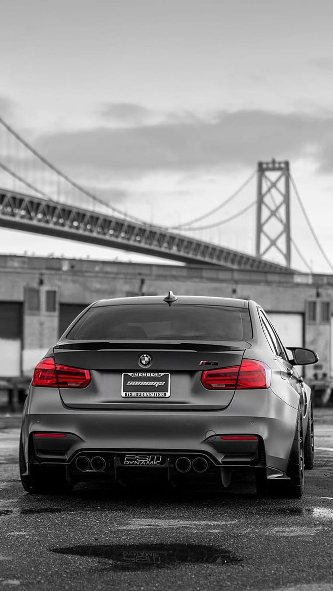 Matte black BMW M3 rear view parked on wet pavement with the San Francisco Bay Bridge stretching into the distance.