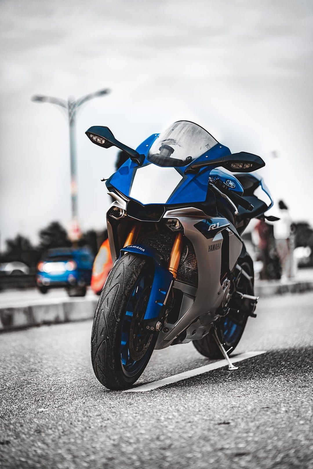Blue Yamaha R1 sportbike with gold front forks parked on a textured asphalt road with a blurred urban backdrop.