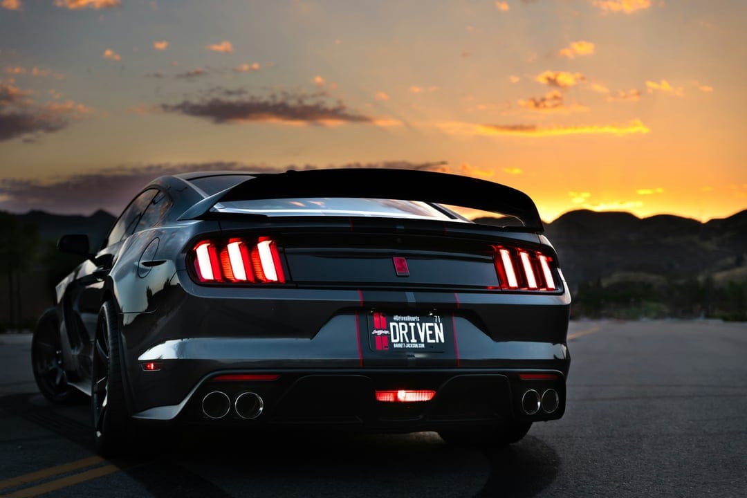Black Ford Mustang GT with glowing red taillights parked on a highway during a dramatic mountain sunset.
