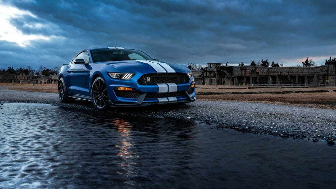 Blue Ford Mustang Shelby GT350R with white stripes on a wet gravel path under a dramatic overcast sky.