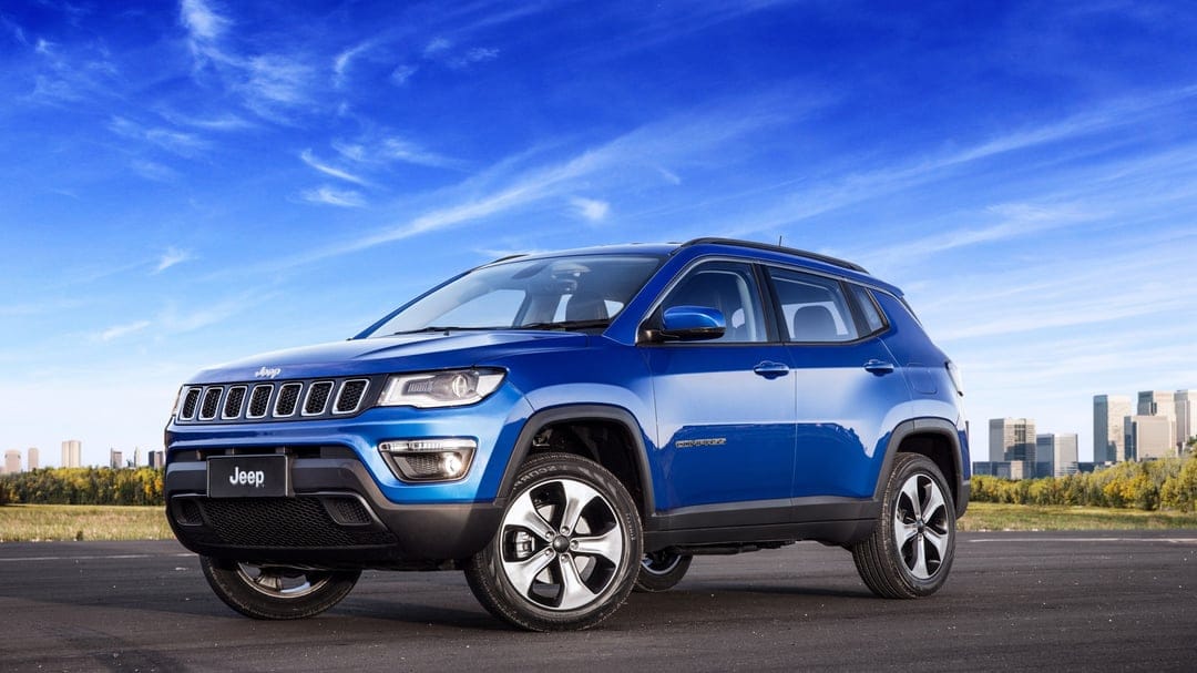 Blue Jeep Compass SUV parked on asphalt under a bright sky with a distant city skyline and trees.