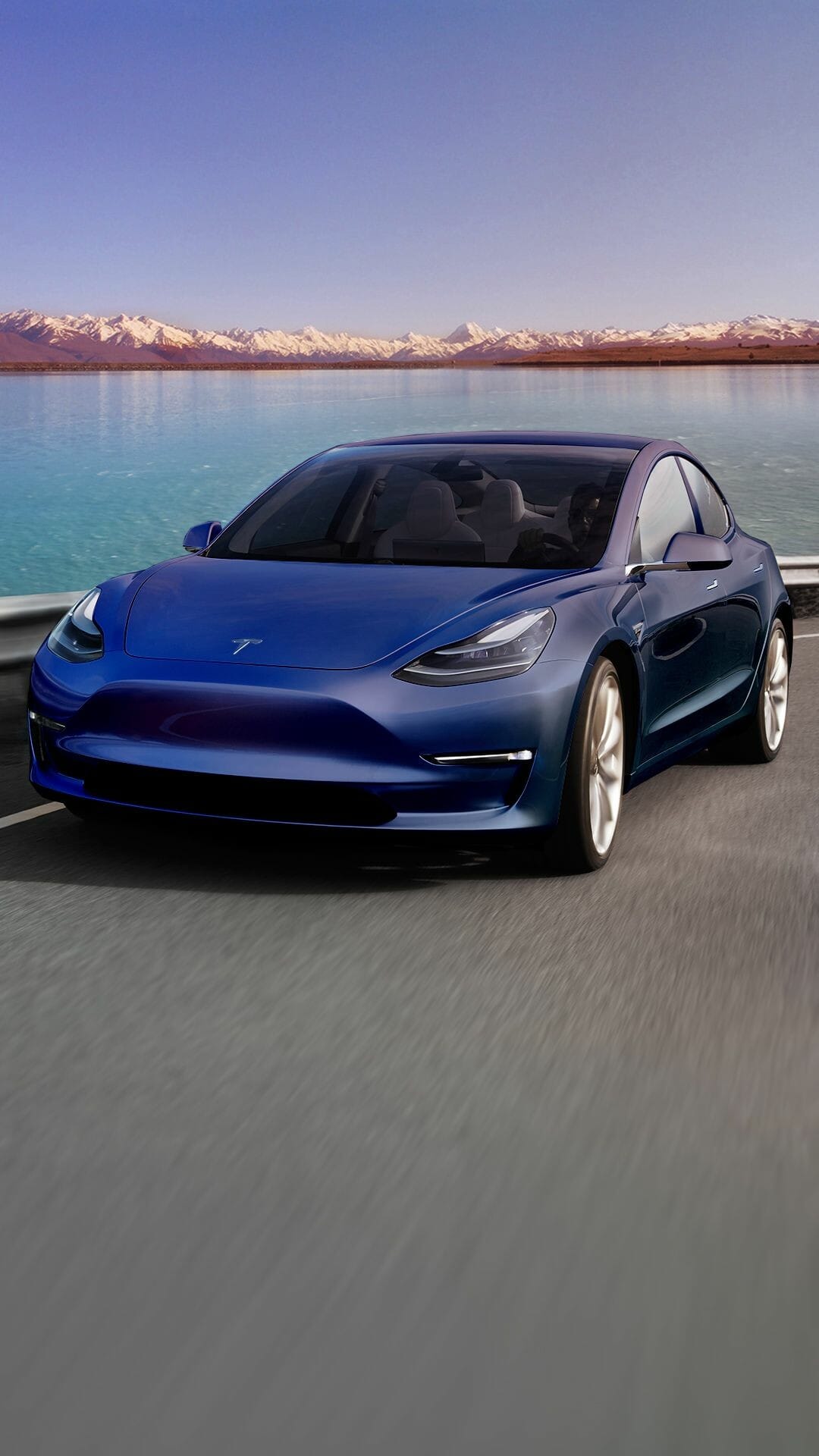 Blue Tesla Model 3 driving on a road beside a lake with snow-capped mountains in the background.