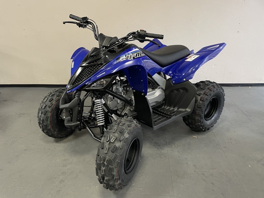 Blue Yamaha Raptor sport ATV with knobby tires and white suspension springs on a gray studio floor.