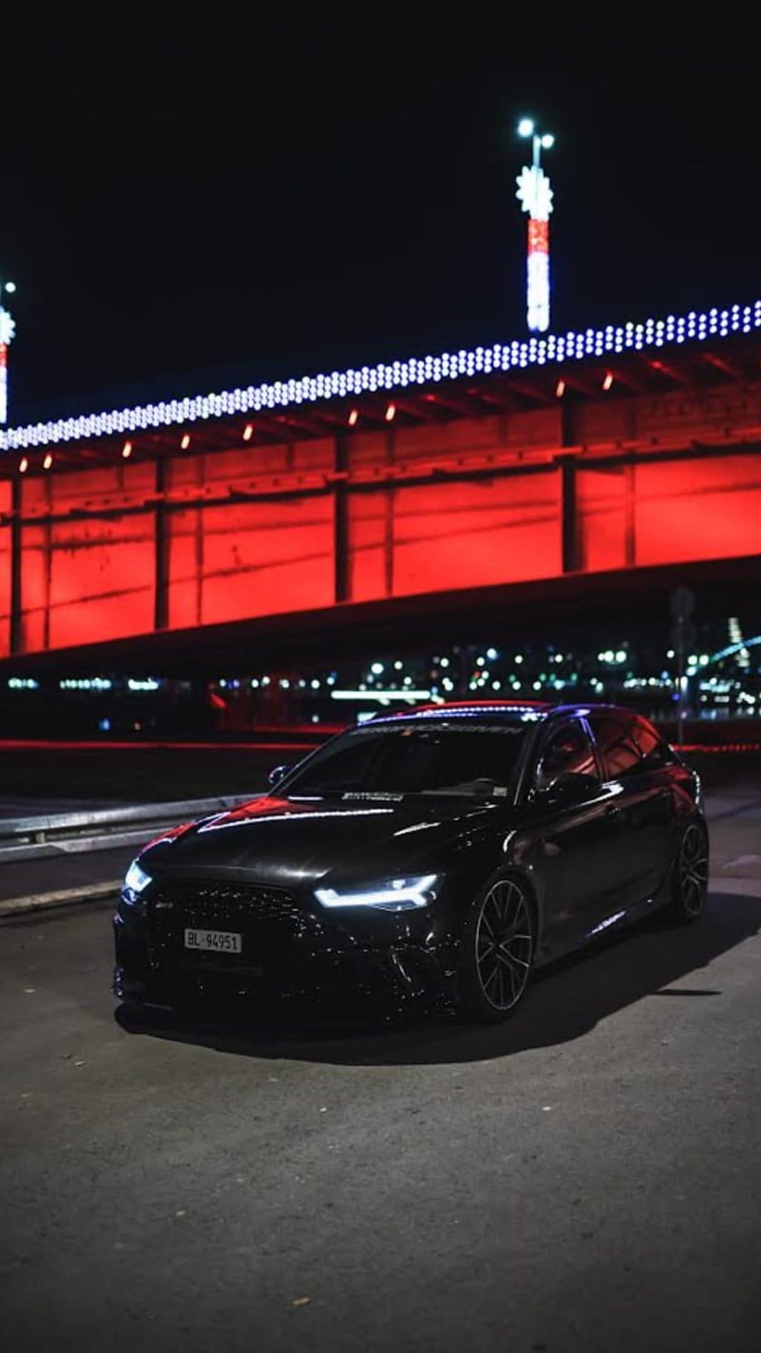 Black Audi RS6 Avant with glowing headlights parked on a city street under a red illuminated bridge at night.