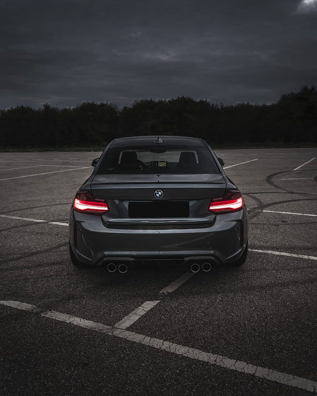 BMW M2 Coupe rear view with illuminated red taillights on a dark asphalt lot under a cloudy twilight sky.