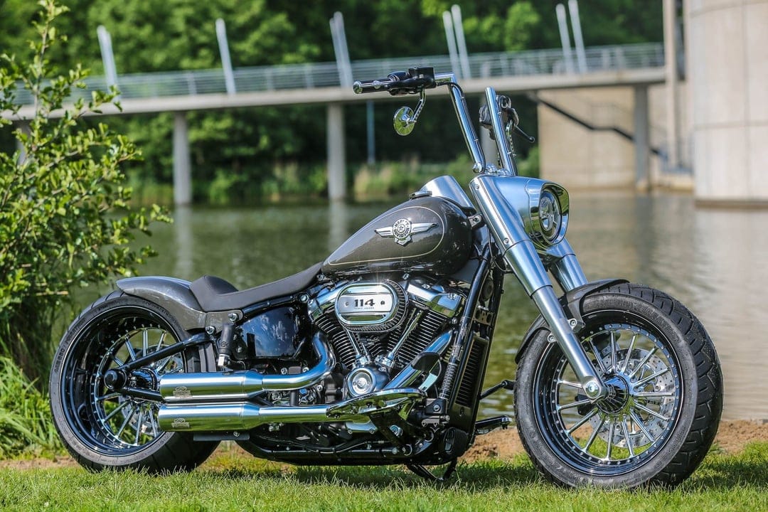 Custom Harley-Davidson Fat Boy 114 motorcycle with chrome accents parked on grass by a lake and a bridge.