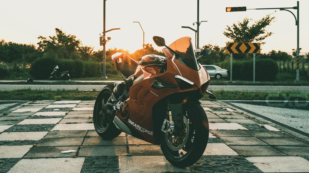 Red Ducati motorcycle parked on a checkered tile pavement during golden hour with a soft city sunset background.