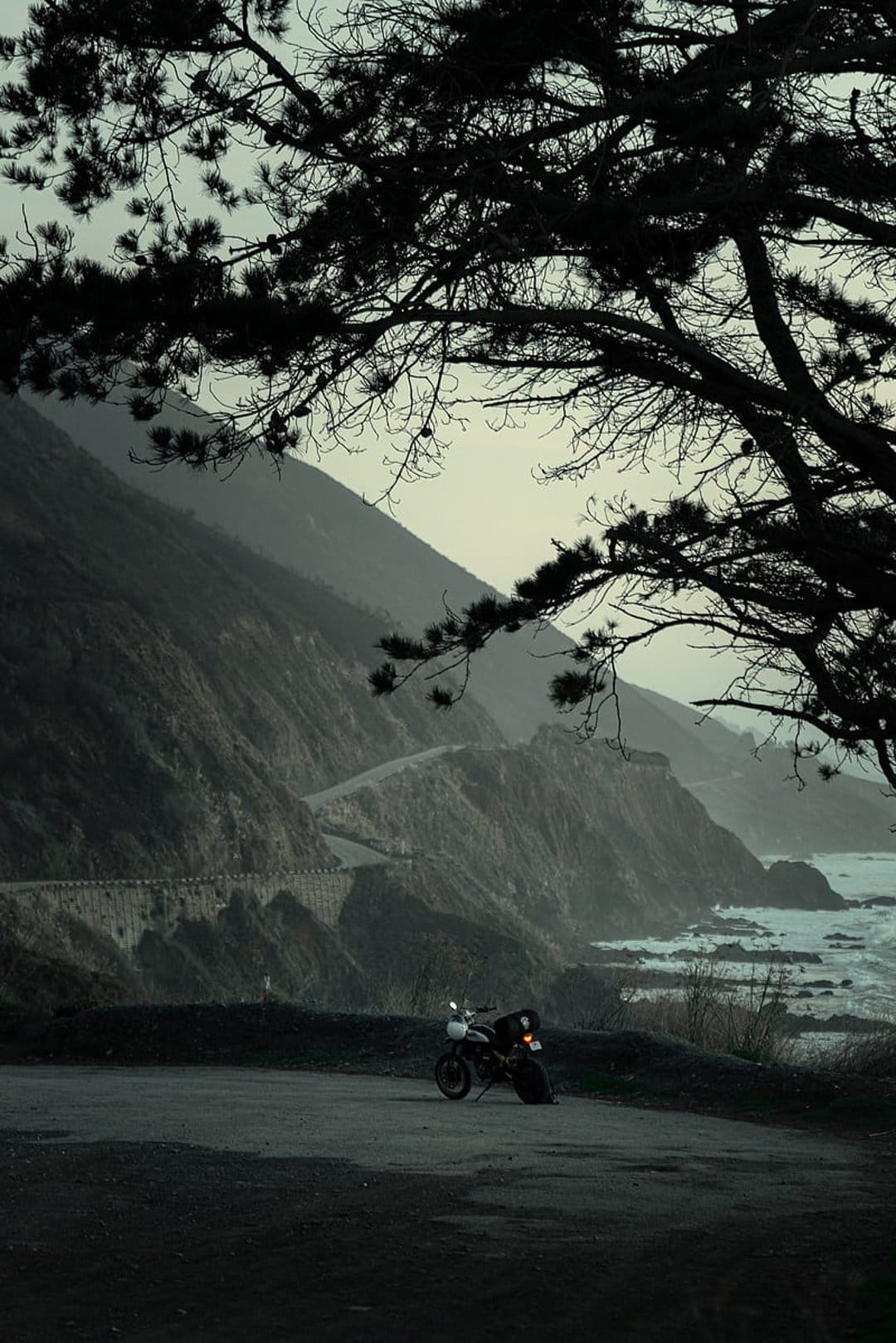 Vintage motorcycle parked on a foggy coastal cliffside road framed by dark tree branches and ocean waves.