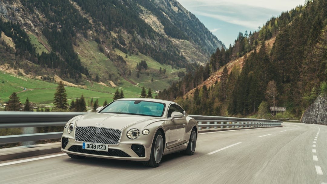 Champagne Bentley Continental GT on a winding mountain road with green slopes and forests in the background.