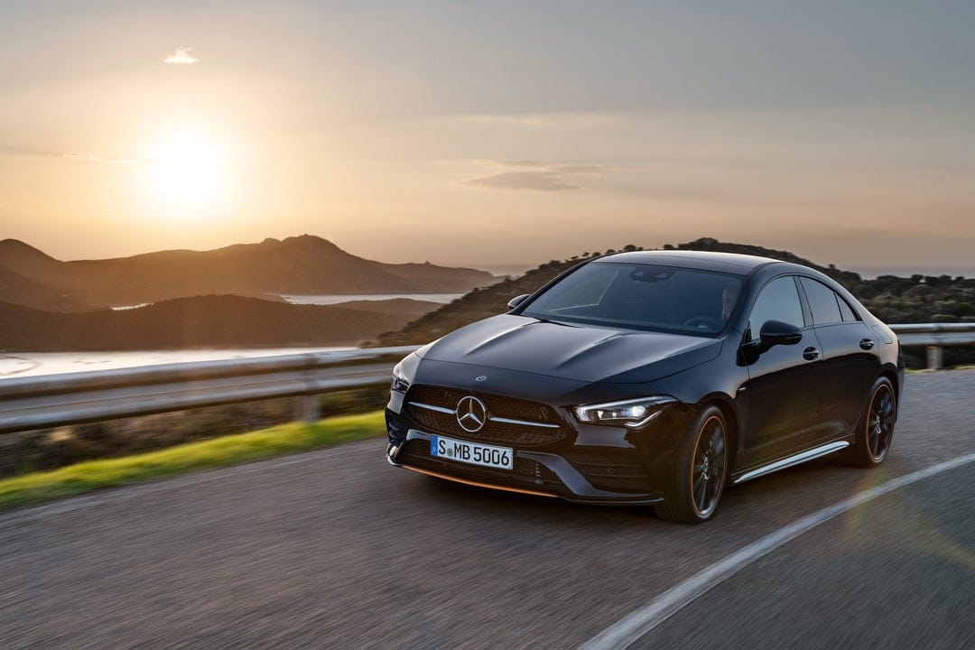 Black Mercedes-Benz CLA driving on a coastal road at sunset with golden lighting and mountain scenery.