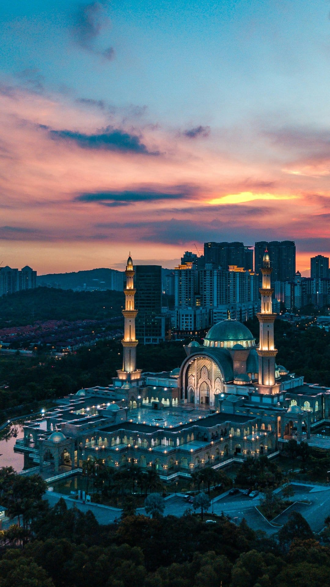 Grand mosque minarets reflect in water beside a metropolitan skyline during a vibrant orange and purple sunset.