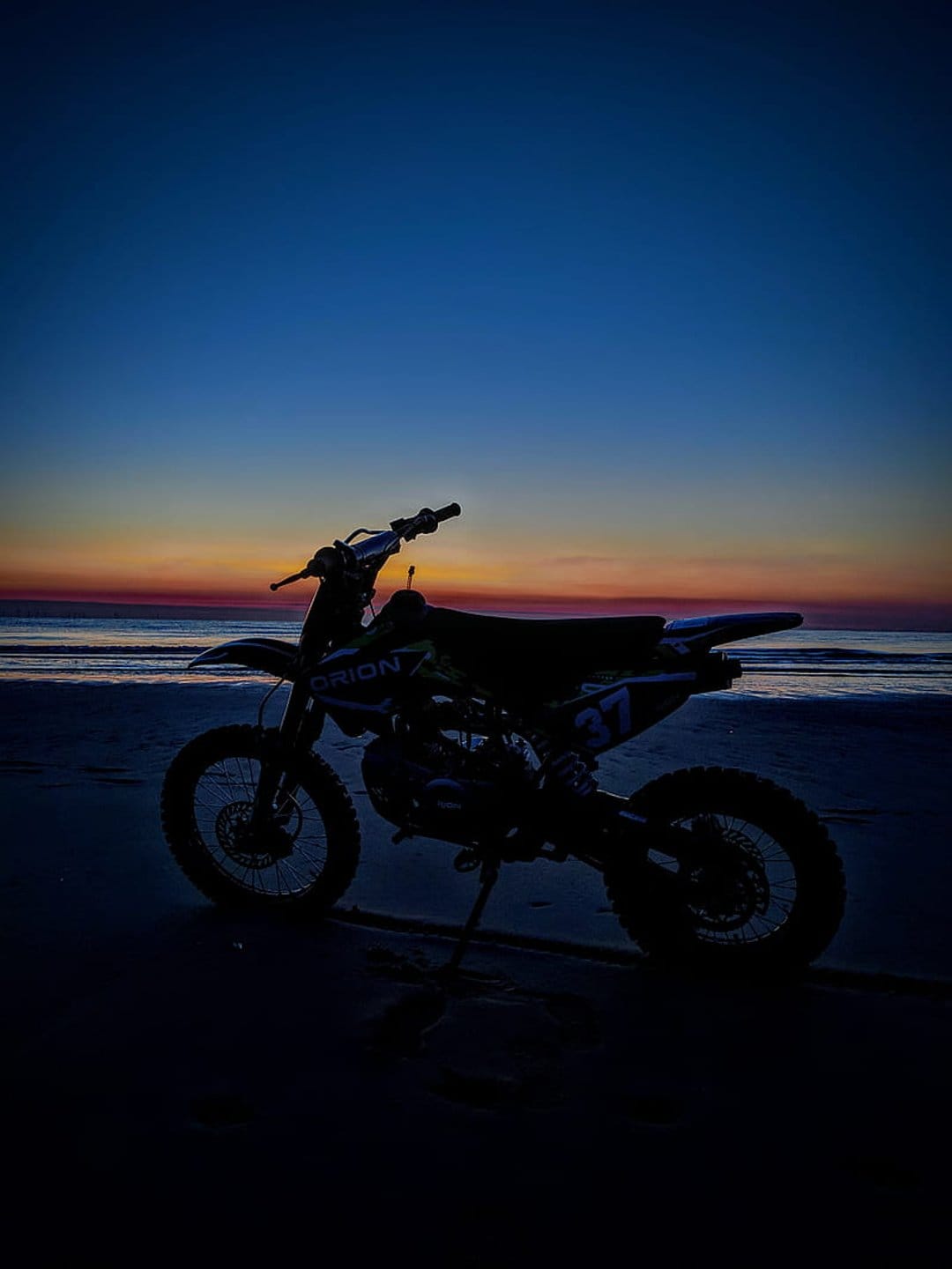 Dirt bike silhouette on a sandy beach against a vibrant orange and blue sunset sky with distant ocean waves.
