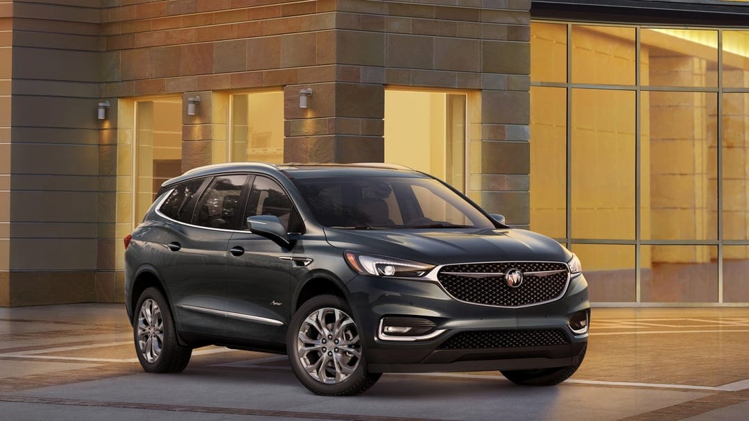 Dark green Buick SUV parked in front of a modern stone building with glowing golden windows during the evening.