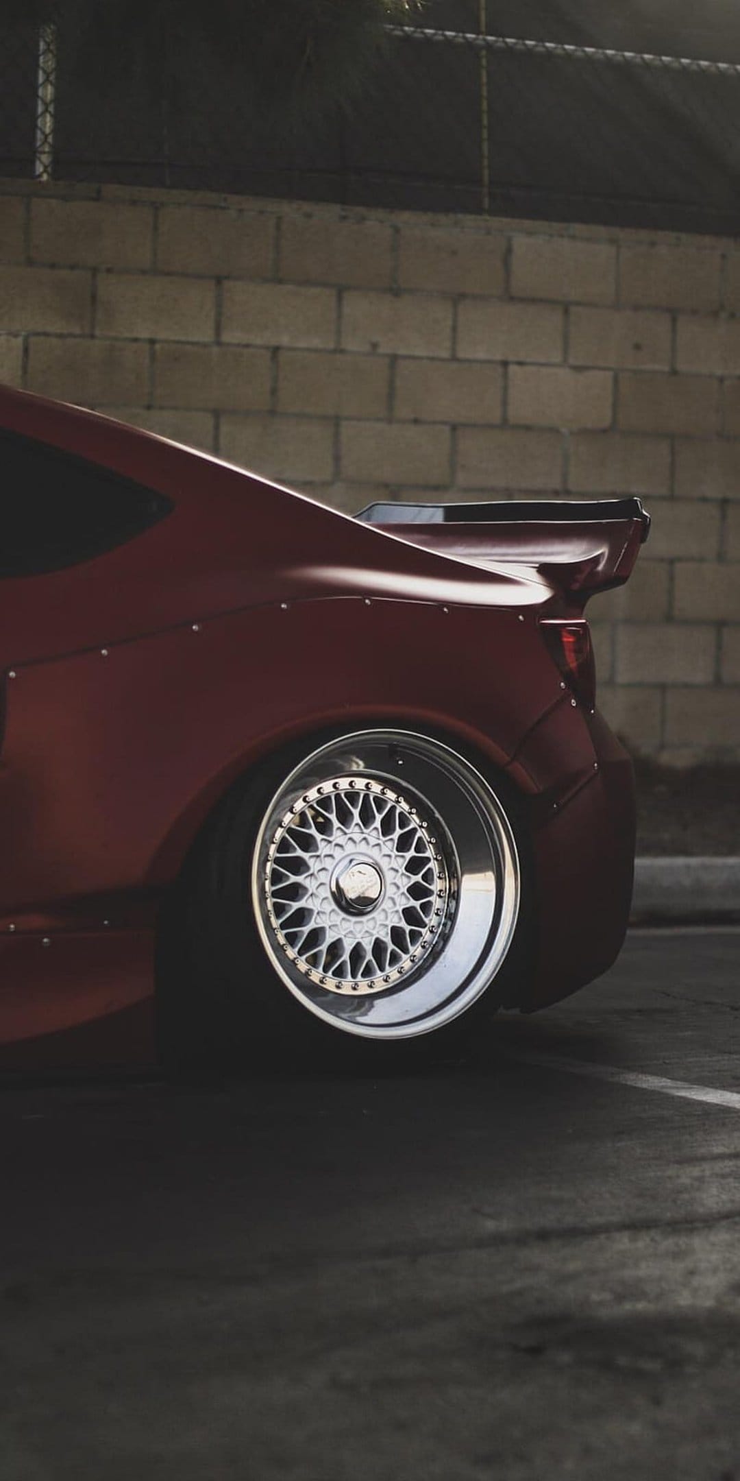 Red widebody sports car with chrome mesh wheels and a tall rear wing parked against a textured brick wall.