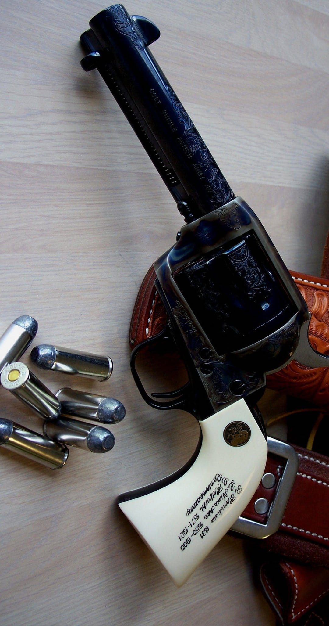 Engraved Colt Single Action Army revolver with ivory grips on a wooden surface next to leather gear and bullets.