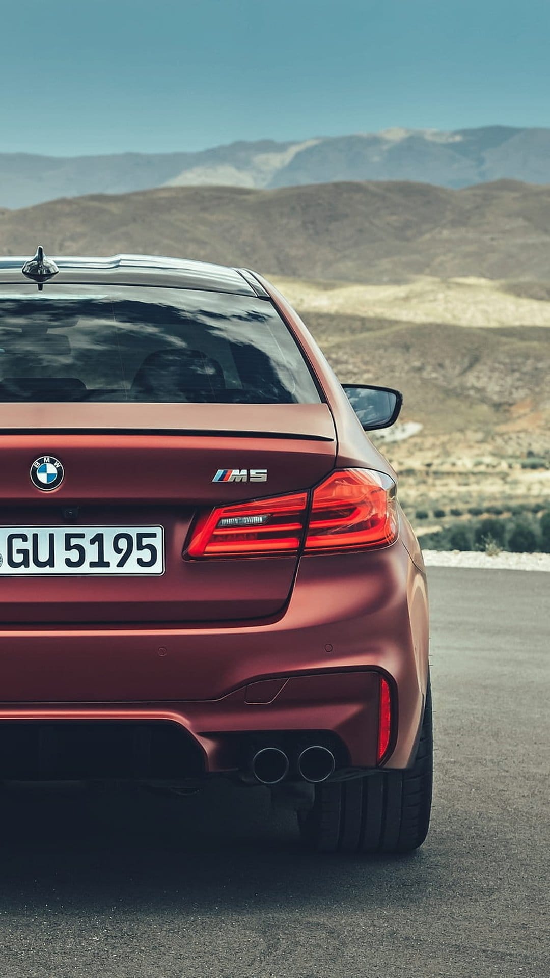 BMW M5 red rear bumper, quad exhausts, and taillights on a desert asphalt road with distant hazy mountains.
