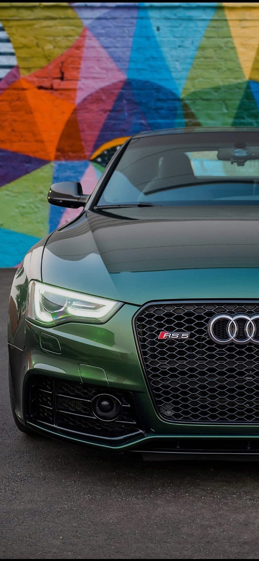 Green Audi RS5 coupe front view against a brick wall with colorful graffiti art in an urban street setting.