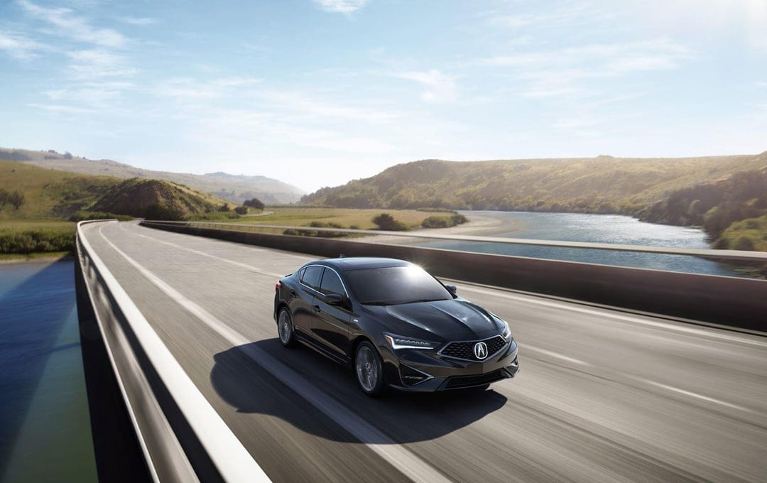 Black luxury sedan driving on a curved bridge over a river with hills and blue sky in the background.