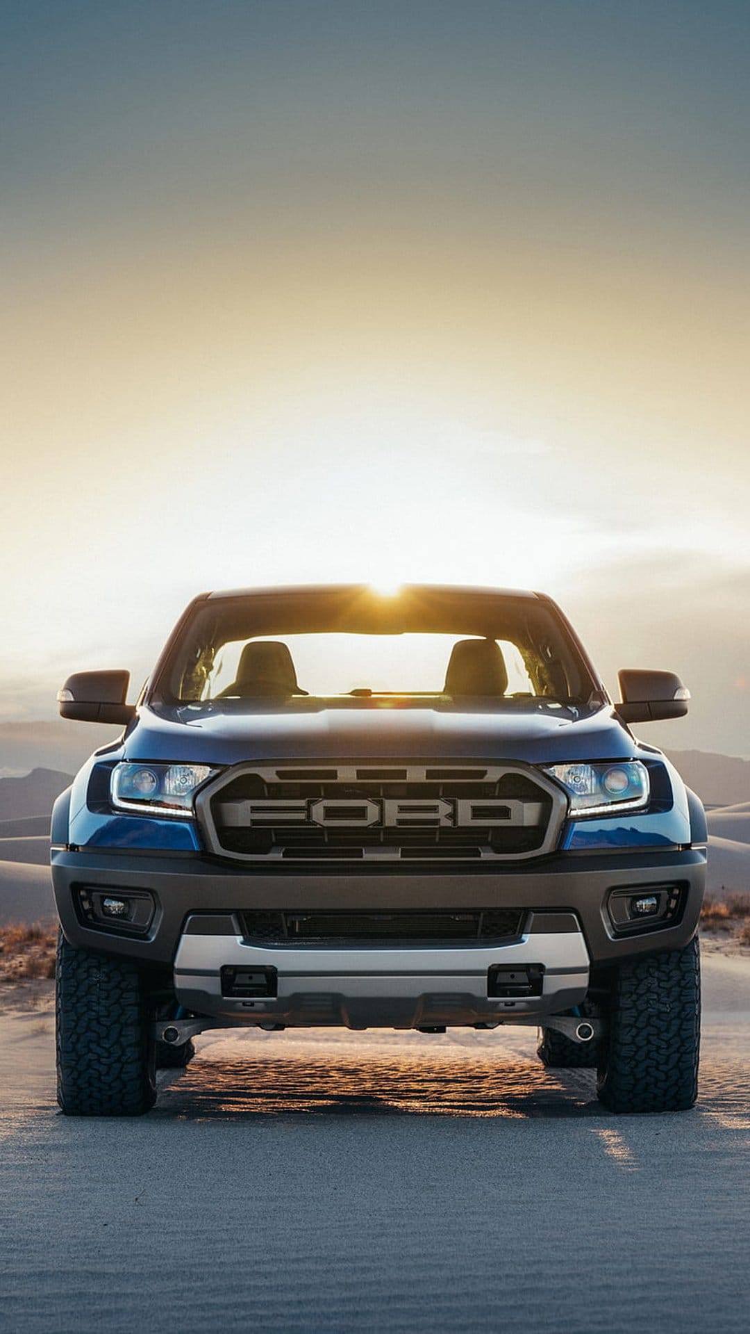 Blue Ford Ranger Raptor truck driving through desert sand dunes during a golden sunset with a prominent grille.