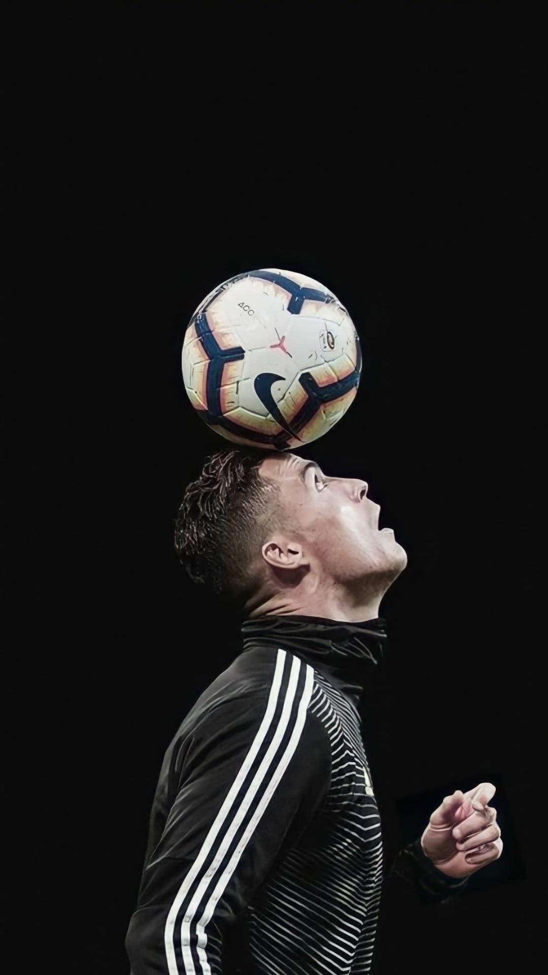Cristiano Ronaldo balances a soccer ball on his head wearing a black Adidas tracksuit against a dark background.