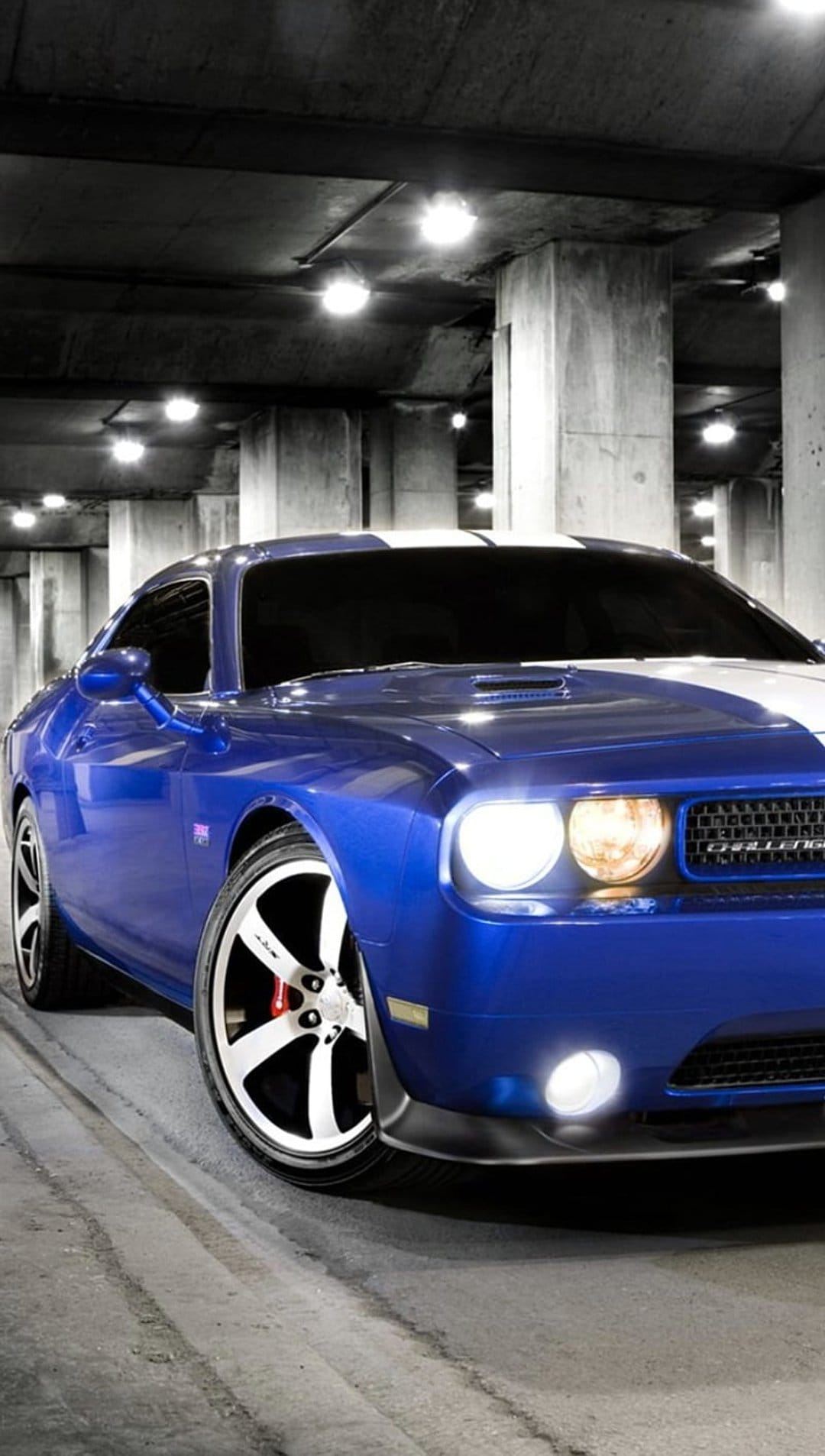 Blue Dodge Challenger with white stripes and glowing headlights parked in a concrete industrial garage.