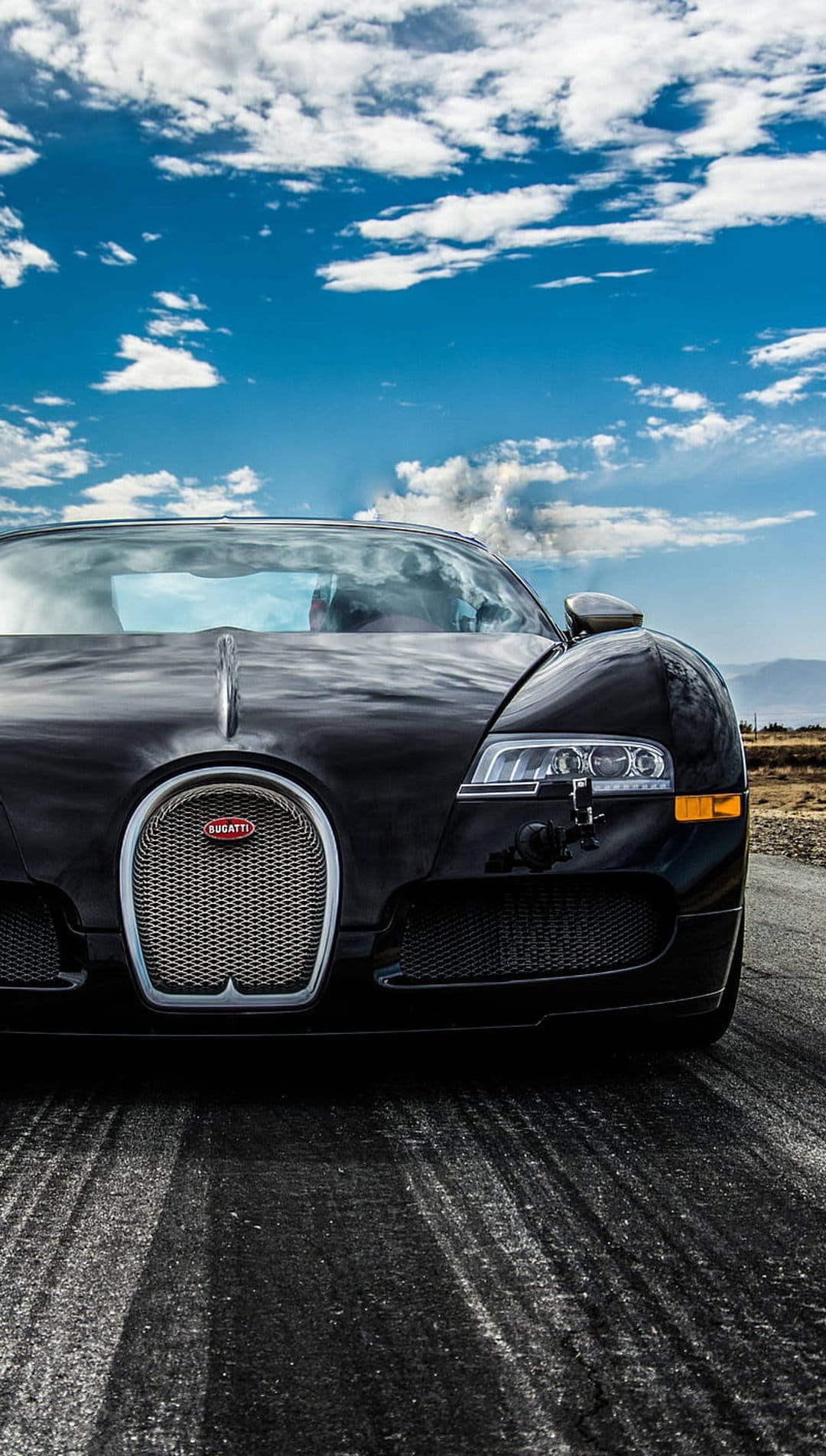 Black Bugatti Veyron supercar on an asphalt road under a cloudy blue sky with mountains in the background.