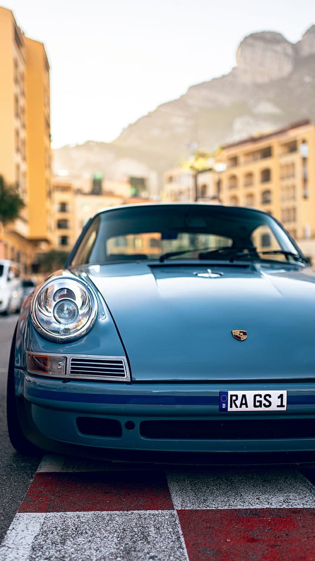 Blue Porsche 911 classic car parked on a Monaco street with Mediterranean buildings and mountains behind it.