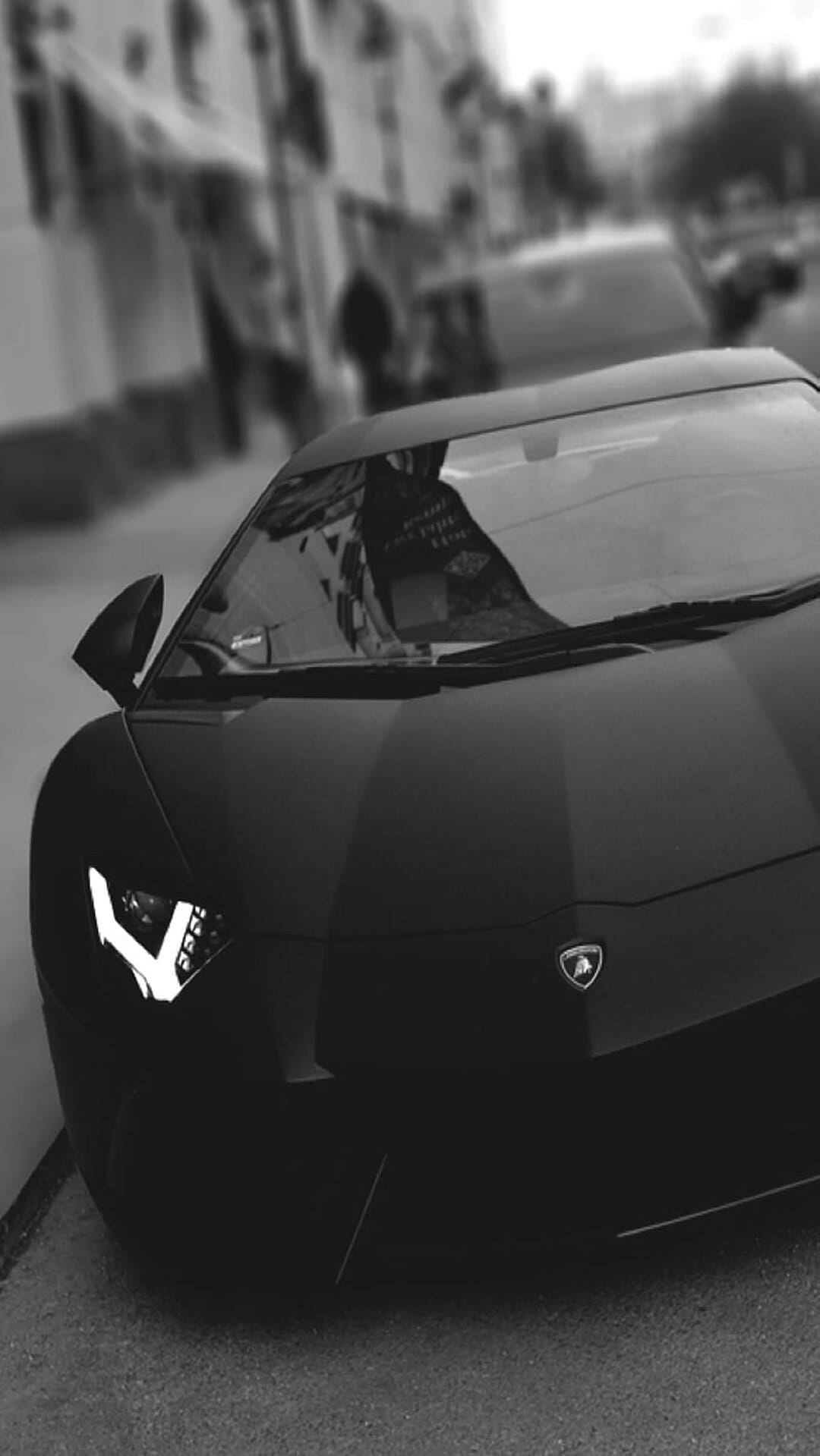 Black Lamborghini Aventador with glowing Y-shaped headlights on a city street in a vertical monochrome shot.