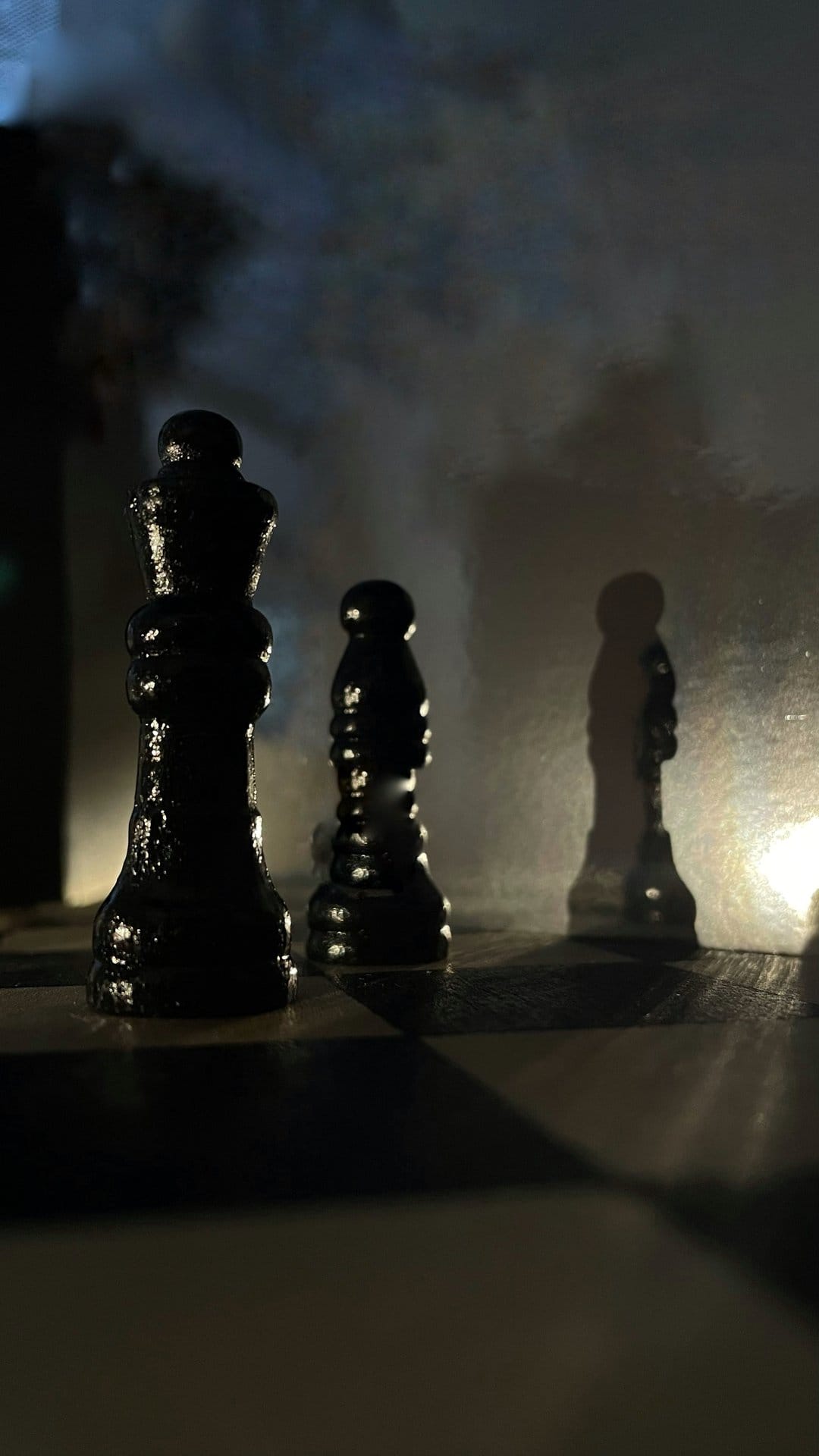 Black chess queen and pawn pieces standing on a board amidst thick atmospheric grey smoke and dark shadows.