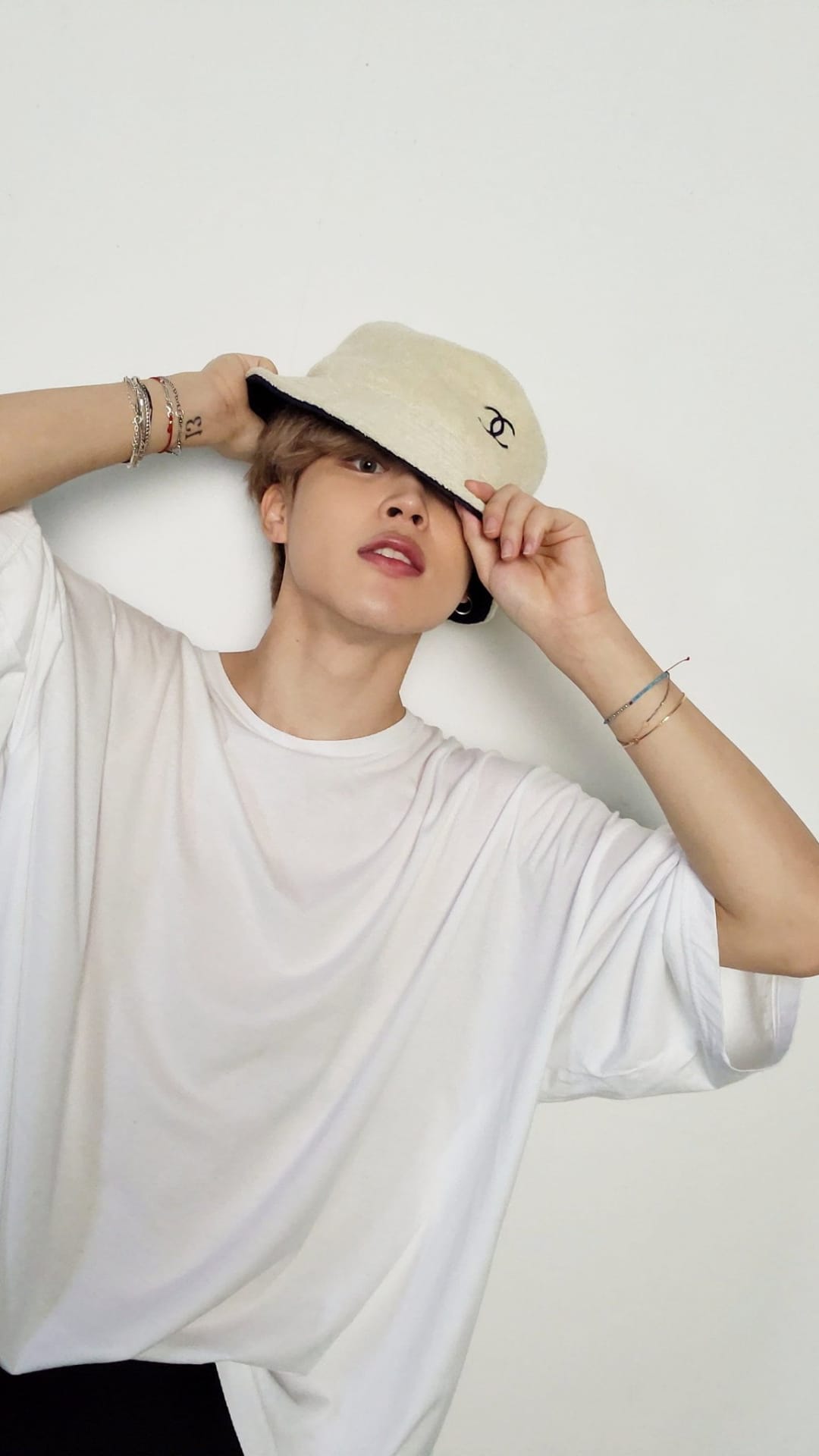 Young man in a white t-shirt and Chanel bucket hat poses against a white wall with his hand on his head.
