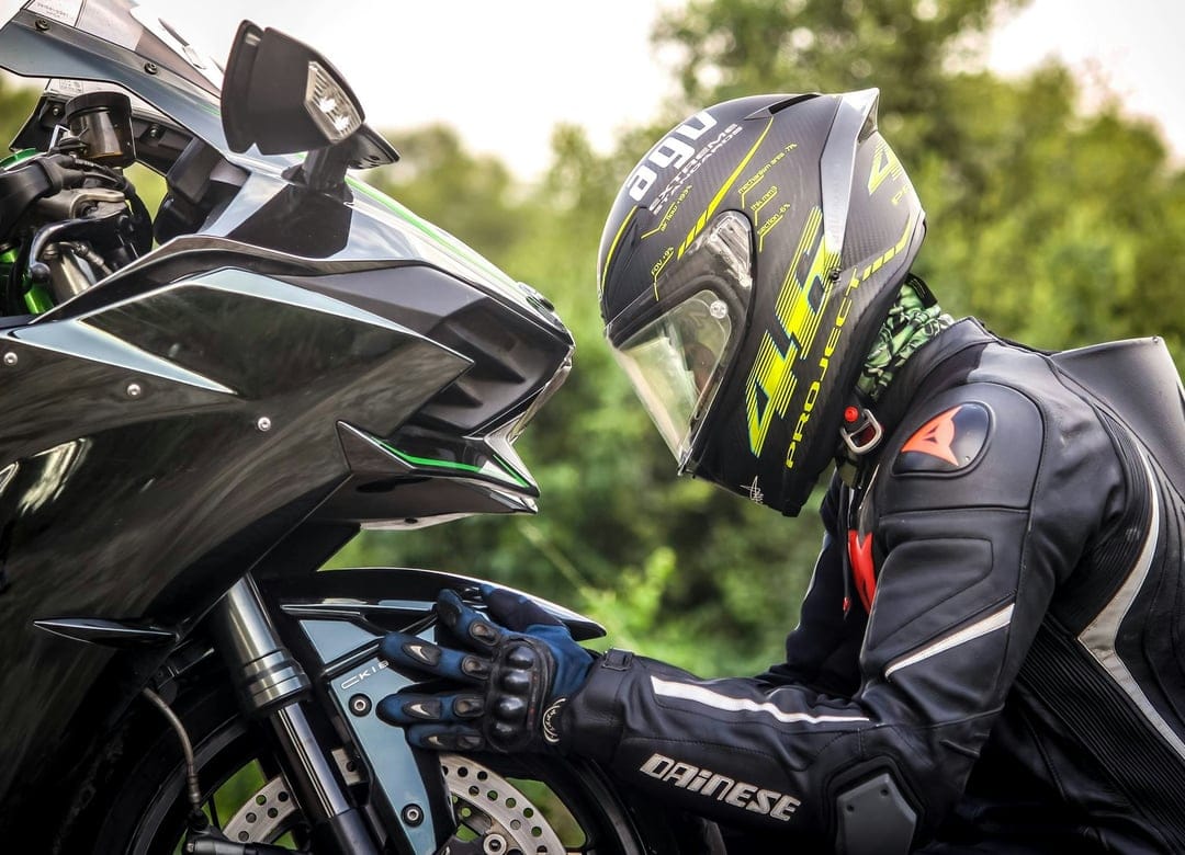 Motorcyclist in a black Dainese racing suit and AGV helmet leans toward a black sportbike with green accents.