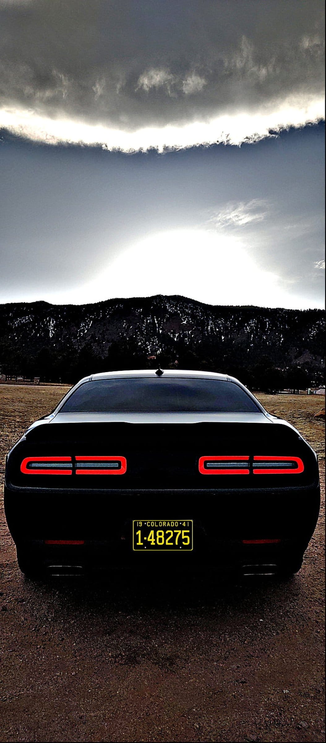 Black Dodge Challenger rear with glowing red LED tail lights and a yellow license plate under a cloudy mountain sky.