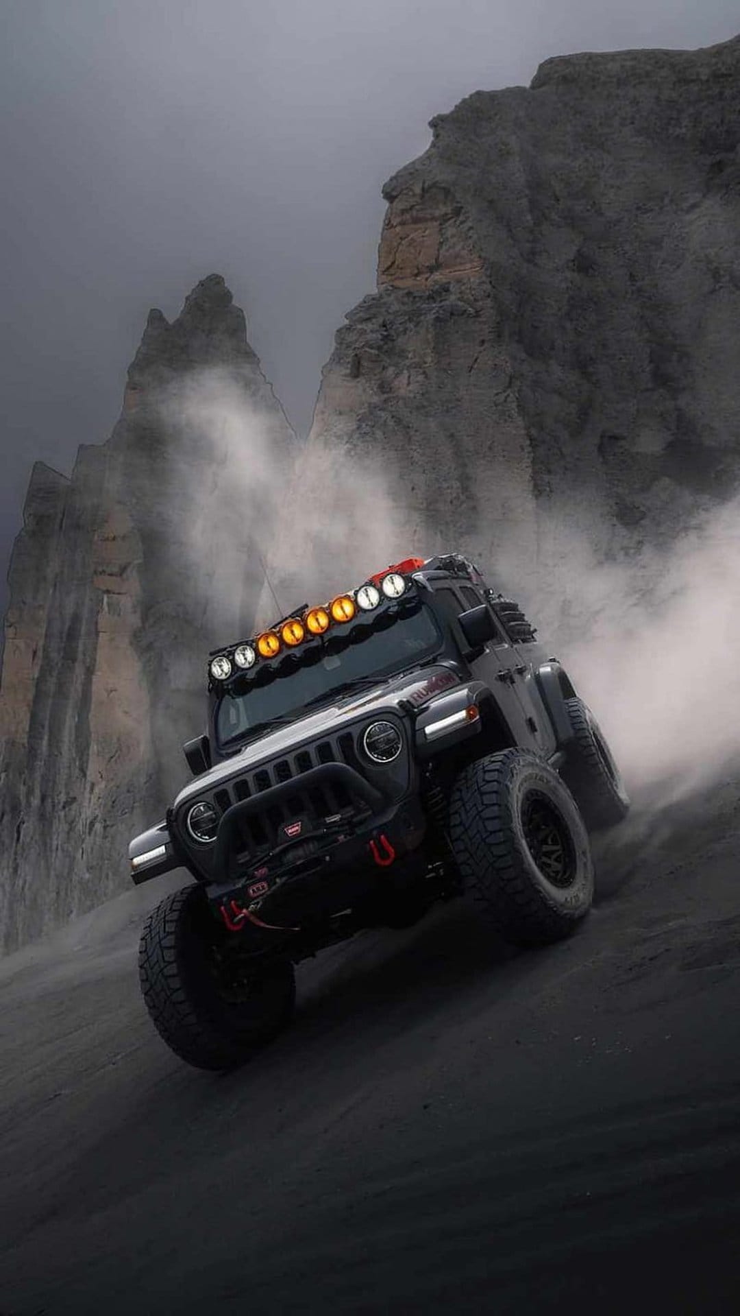 Black Jeep Wrangler climbing a steep dusty trail with bright roof lights against misty mountain rock formations.
