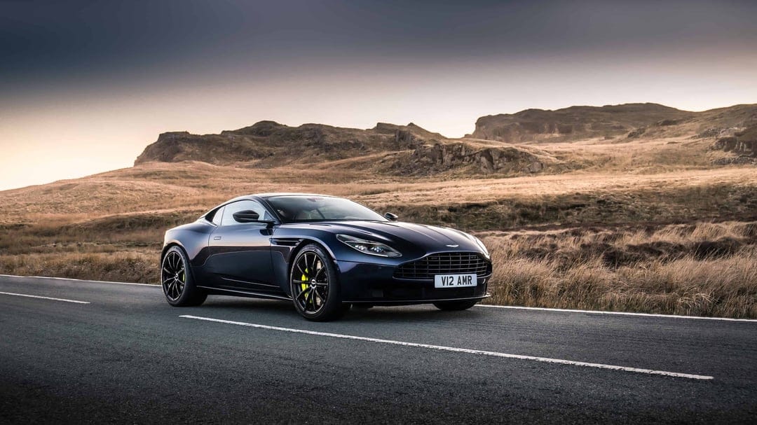 Dark blue Aston Martin DB11 AMR on a paved mountain road with yellow brake calipers and a sunset sky.