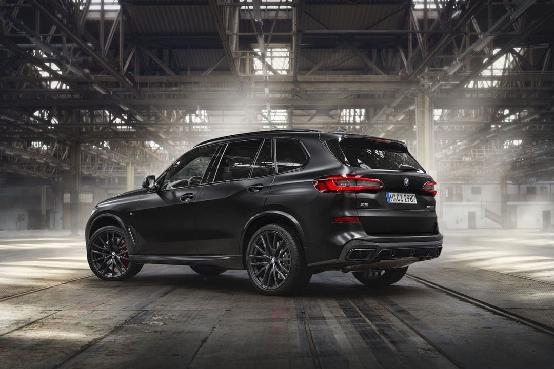 Matte black BMW X5 SUV parked in a sunlit industrial warehouse with dramatic overhead lighting and steel beams.